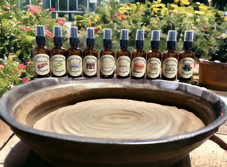 Numerous 2oz spray bottles containing various fragrances of room, spray sitting on a potters wheel with a garden and greenhouse in the background.