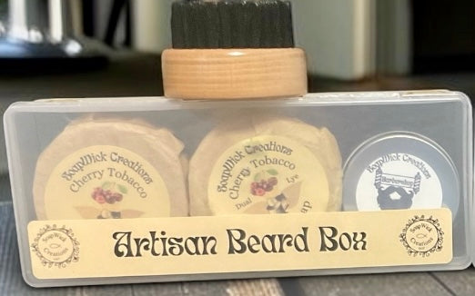 Artisan Beard Box set in a clear plastic case with a wooden-handled brush on top, two round cherry tobacco shave soaps, and a small round tin, labeled “Barbershop Beard Balm”.