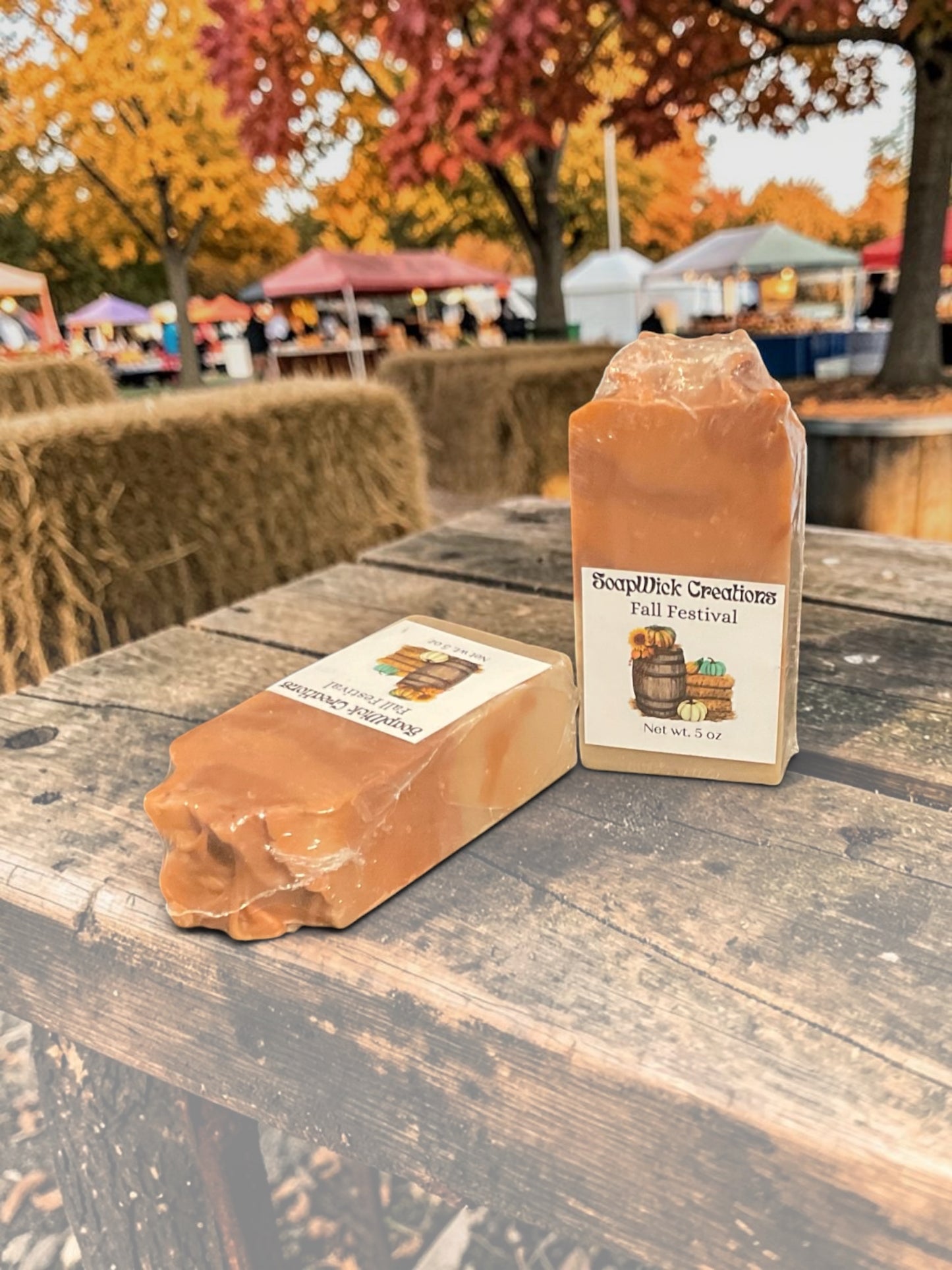 Two bars of fall festival soap, sitting on a vendors table with hay bales and a fall festival going on in the background.