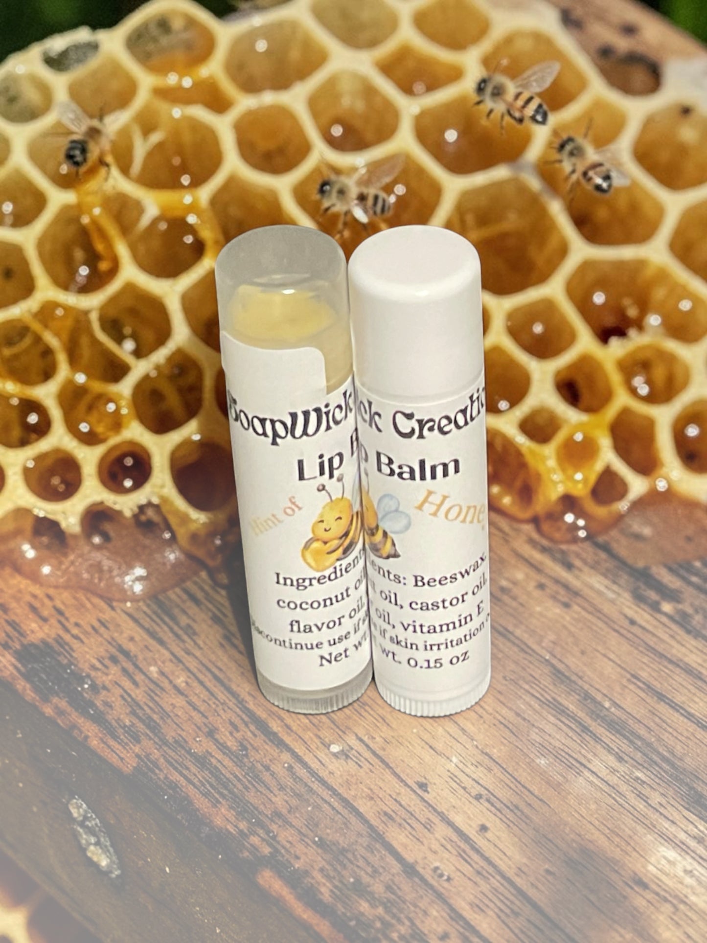 Two plastic lip balm tubes, labeled SoapWick Creations Hint of Honey Lip Balm sitting on top of a beehive with honey dripping honeycomb and honeybees in the background..