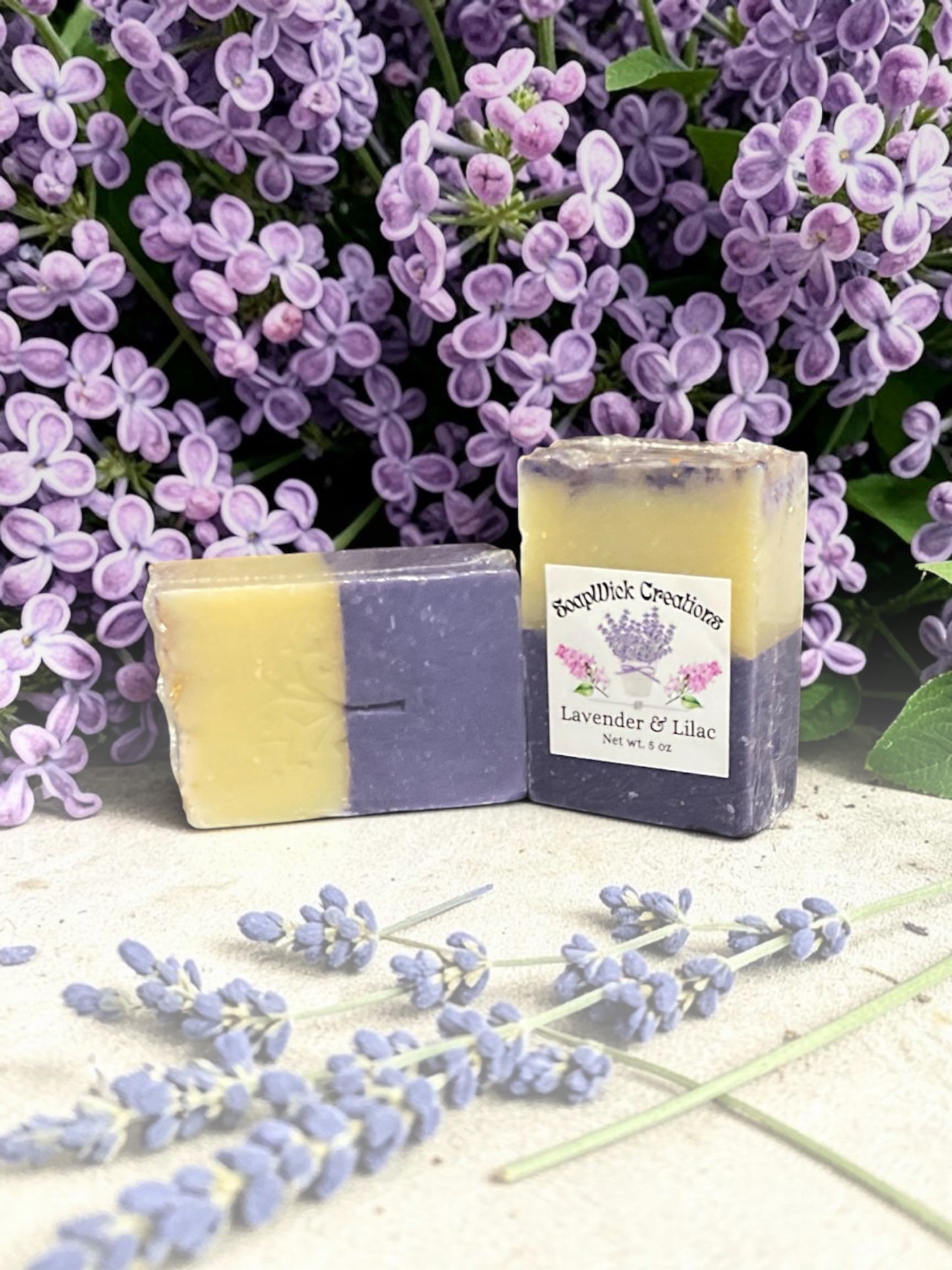 Two Purple and white bar soaps, sitting on a bench with lilac flowers in the background and lavender flowers in the foreground.