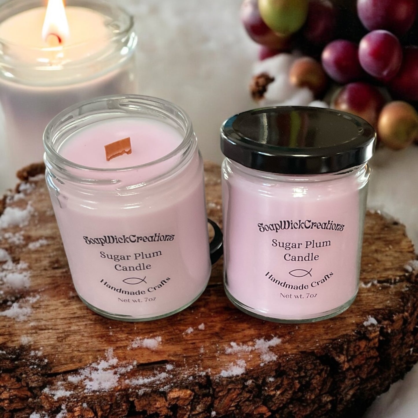 Two pink colored sugar plum scented candles sitting on a slice of log with plums and a candle in the background.