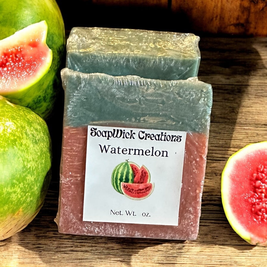 Two pink and green bars of watermelon, scented soap on a wooden table with sliced and whole watermelons.