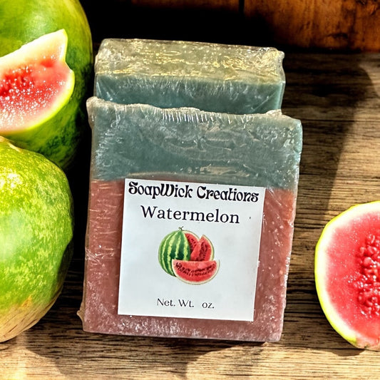 Two pink and green bars of watermelon, scented soap on a wooden table with sliced and whole watermelons.