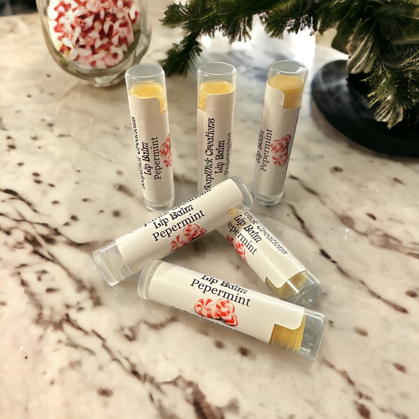 Photograph of six Peppermint flavored lip balm tubes with a Christmas tree and a jar of peppermint candies in the background.