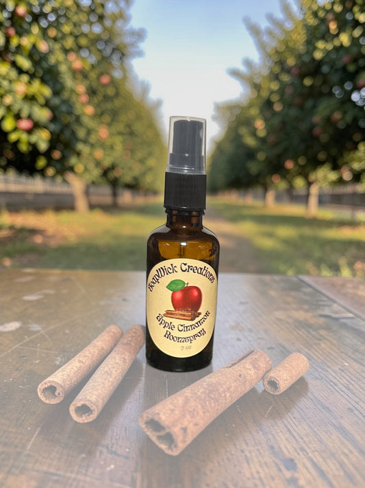 A small amber glass spray bottle labeled 'SoapWick Creations Apple Cinnamon Room Spray.  Sitting on a wooden surface outdoors, with cinnamon sticks in front and blurred apple trees in the background.