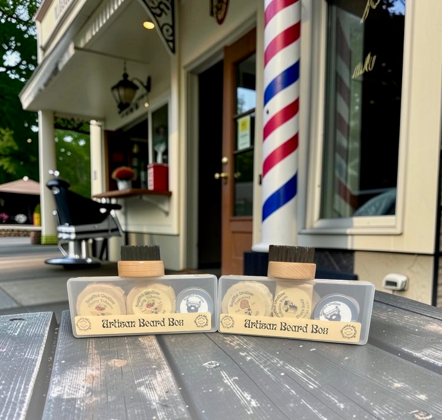 Two boxed beard grooming sets displayed on a wooden table outside a barbershop, with a red, white, and blue barber pole and shop facade blurred in the background.