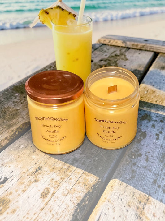 Two orange colored mango and coconut, scented candles, sitting on a wooden table on a beach with a piña colada in the ocean in the background.