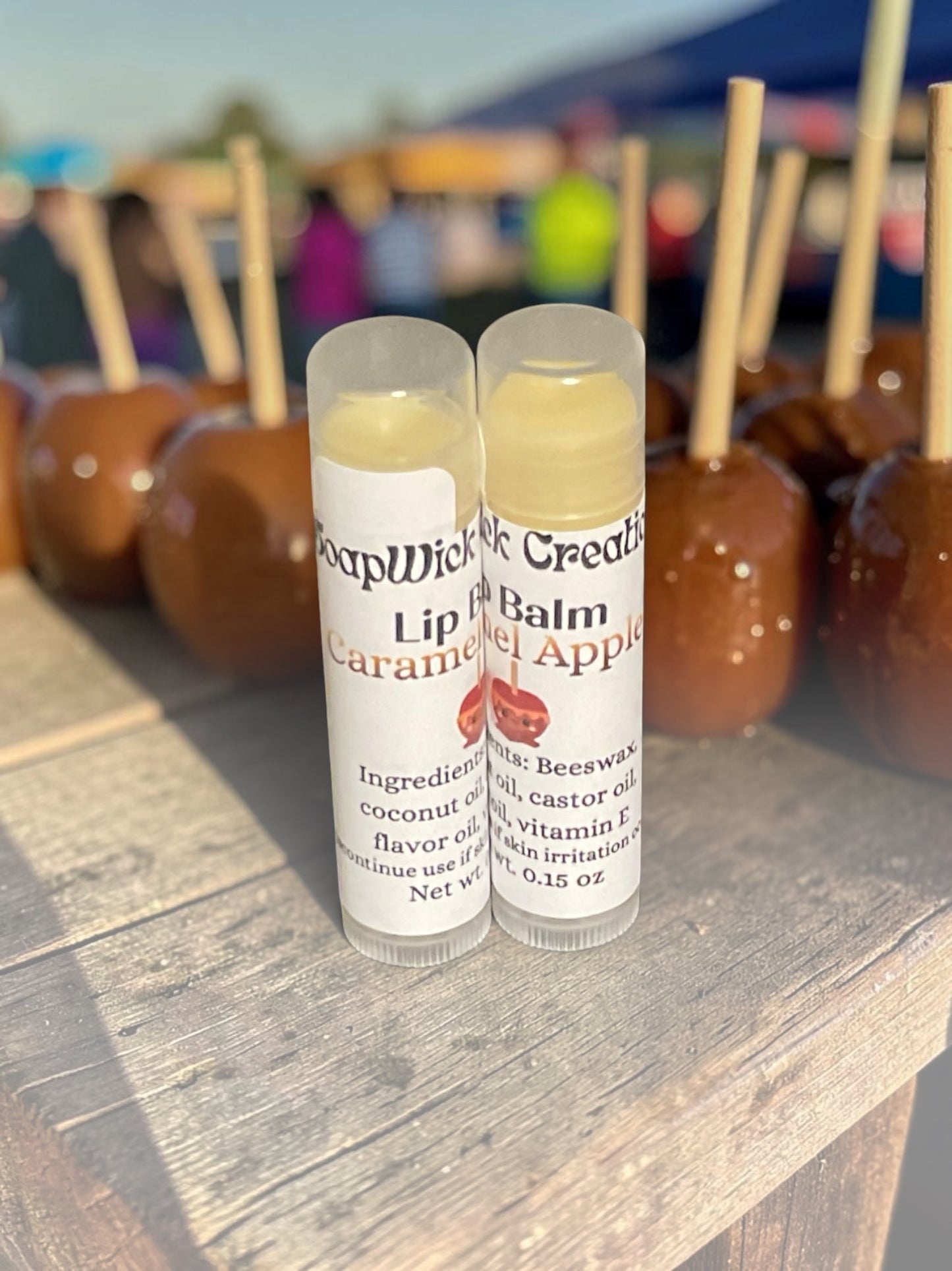 Two clear lip balm tubes labeled SoapWick Creations Caramel Apple flavor, standing on a wooden table with blurred caramel apples in the background.
