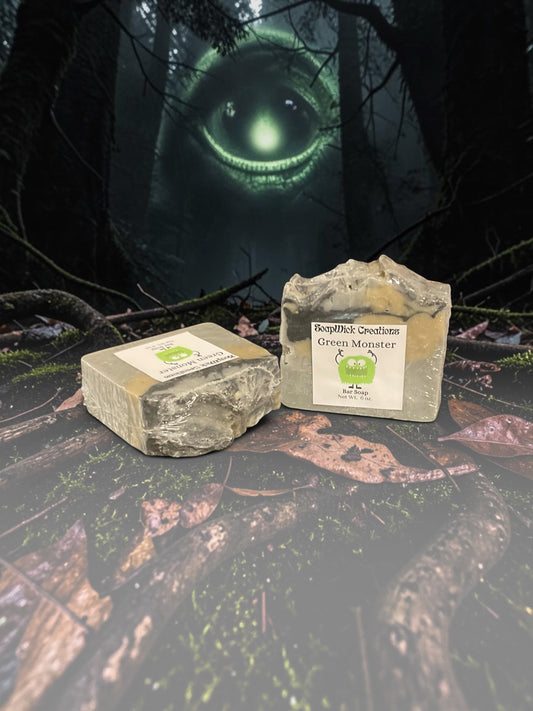 Two bars of beige, black and gold, Green Monster soap sitting on a log in the middle of a forest with a large green monster in the background.