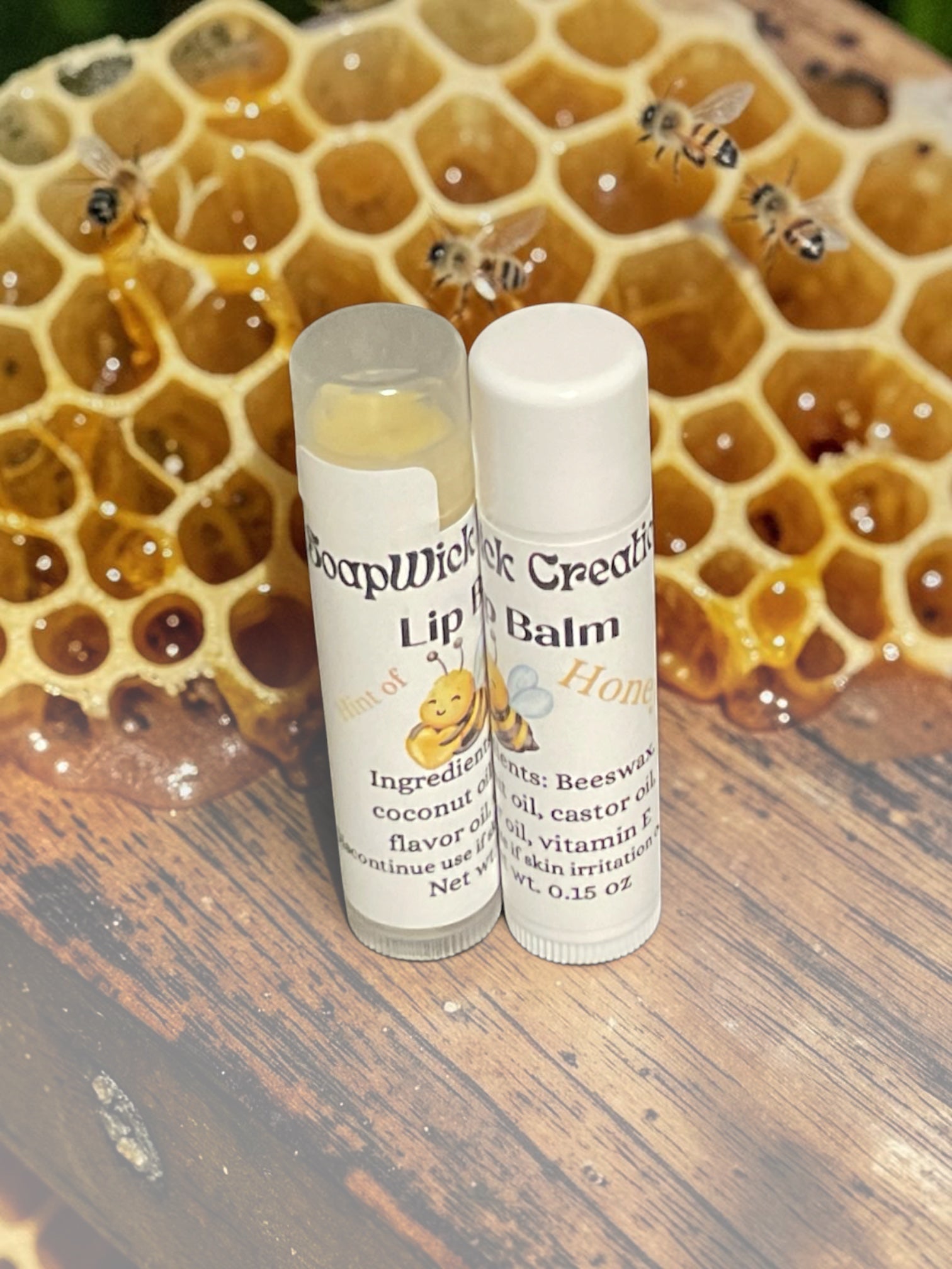 Two plastic lip balm tubes, labeled SoapWick Creations Hint of Honey Lip Balm sitting on top of a beehive with honey dripping honeycomb and honeybees in the background..