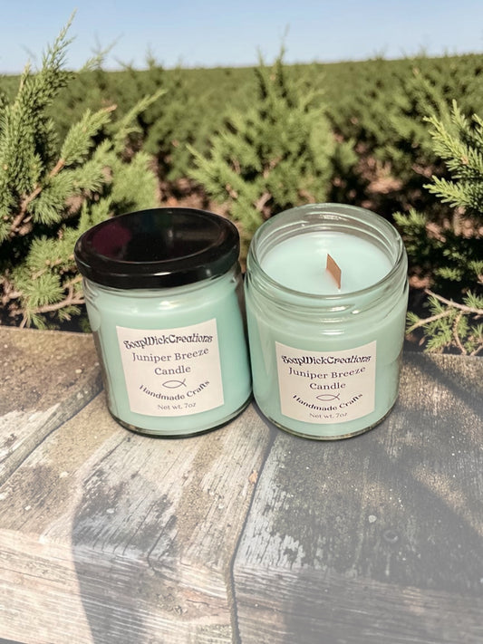 Two light green colored juniper breeze scented candles sitting on a bench, surrounded by spruce trees on a warm breezy day.