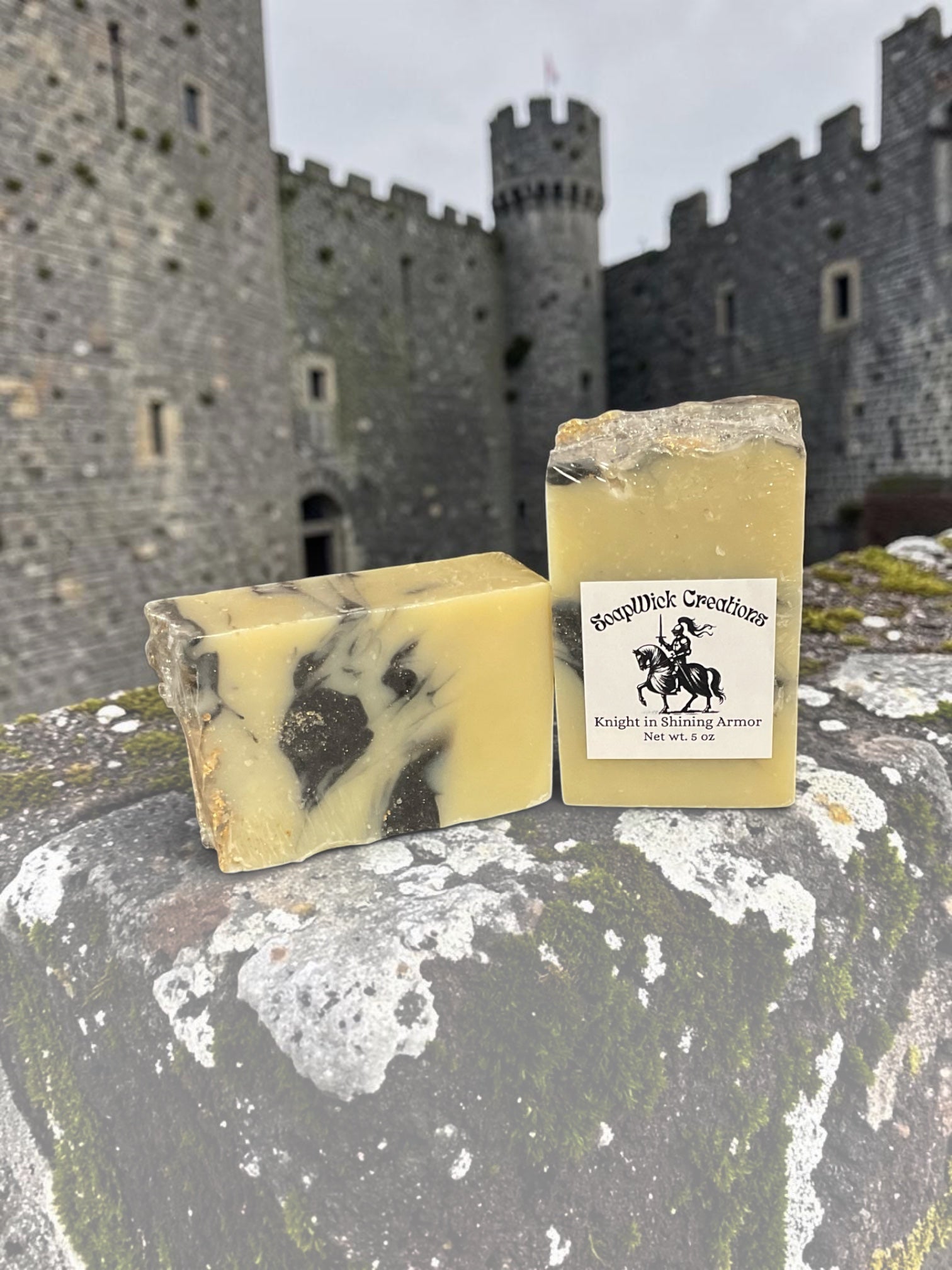 Two cream and black colored bar soaps, sitting on an old stone wall with a medieval castle in the background.