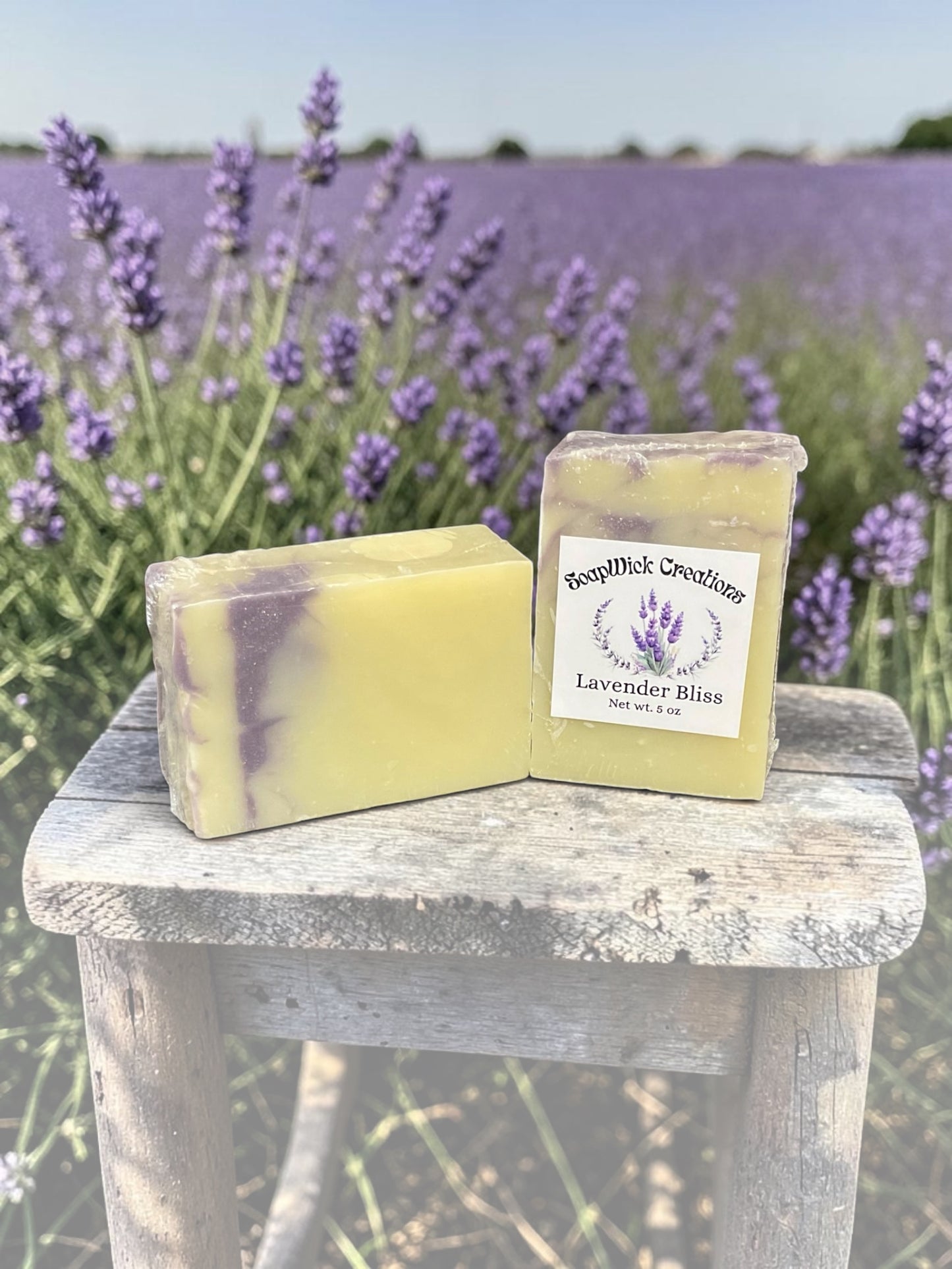 Two lavender scented bar, soaps, purple, and white and color sitting on a bench with a field of lavender behind them.