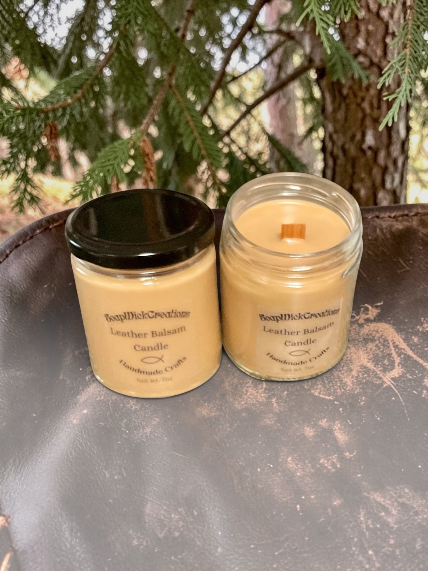 Two beige colored candles with leather balsam scent sitting on a leather saddle in front of a balsam tree.