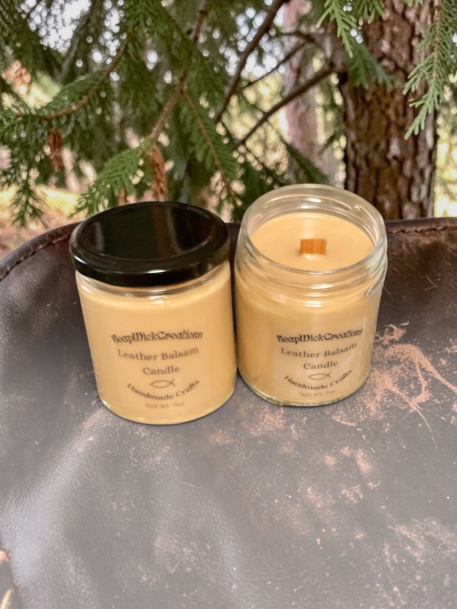 Two beige colored candles with leather balsam scent sitting on a leather saddle in front of a balsam tree.