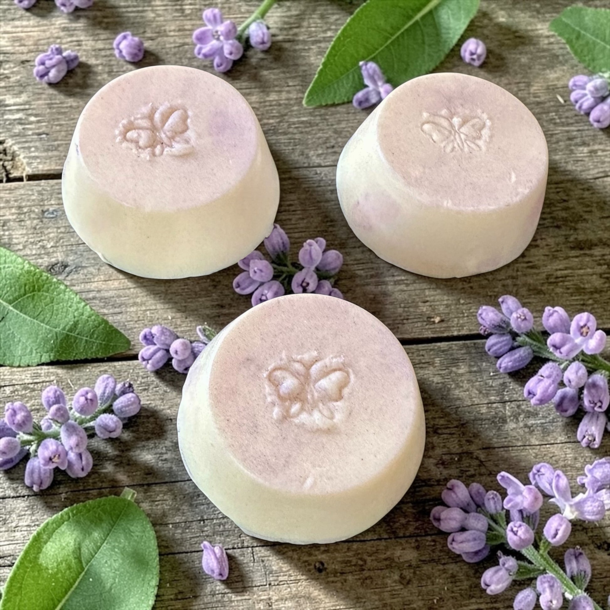 Three lilac scented shampoo bars sitting on a wooden pallet with lilac flowers and leaves in the background.