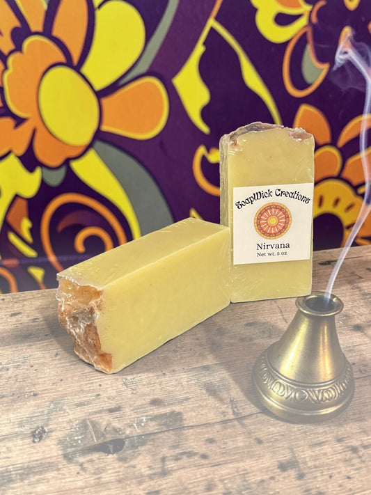 Two beige colored Nirvana bar soaps, sitting on a wooden table with a colorful floral background and an incense burner with incense burning in the foreground.