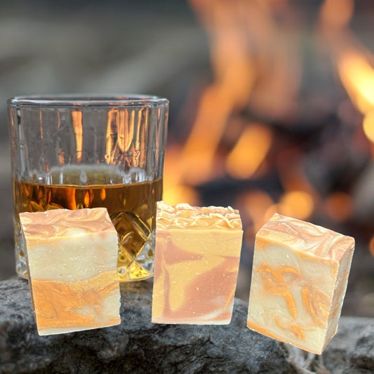 Three soap bars with maple apple bourbon scent with fire and a glass of bourbon in the background .