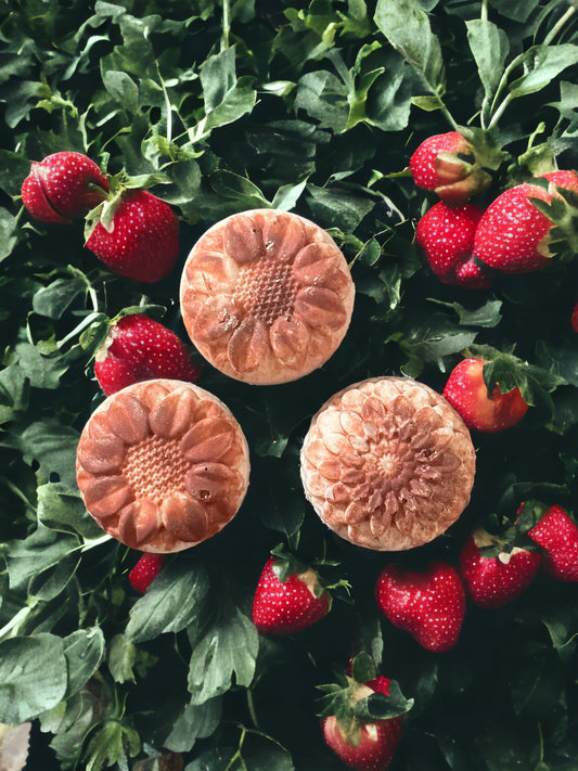 Three strawberry scented shampoo bars with a strawberry bush as a backdrop.