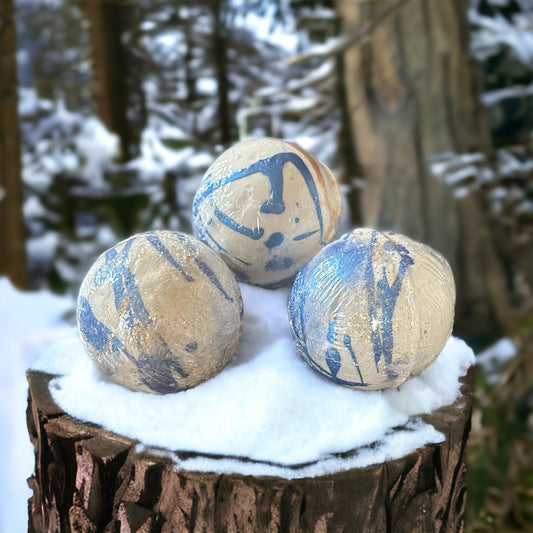 Photograph of three white bath bombs with blue and gold bands on them, sitting on a snow covered stump with forest in the background.