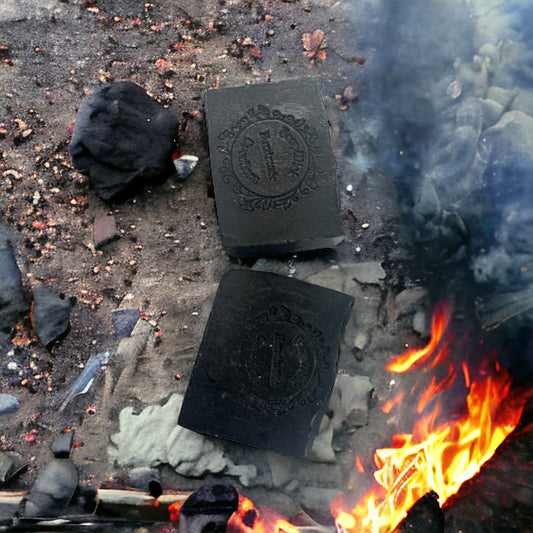 Photograph of two bars of Lump of Coal soap, next to a fire with coal ash and a piece of coal.