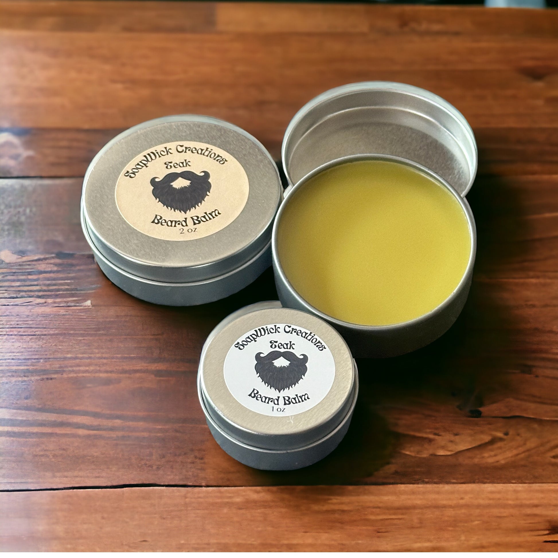 A 1 ounce silver tin and a 2 ounce silver tin containing teak scented beard balm on a wooden table.