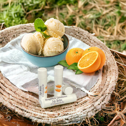 Three lip balm with orange cream, sickle flavoring, sitting on a towel on a woven round pad with a bowl of vanilla ice cream and oranges and Field in the background.