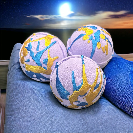 Photograph of three Sleepy Time lavender bath bombs with blue and gold splatter on them sitting on a pillow with a night sky in the background. Scented with Black Chamomile Tea and Lavender scented Fragrance oils.