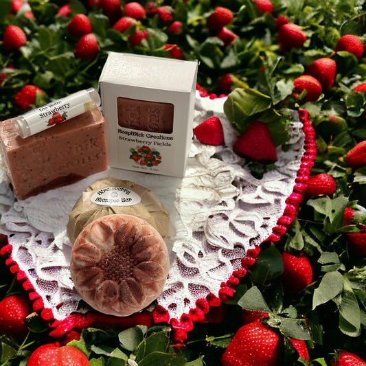 A photograph with white lace heart shaped doily with red trim with two bars of Strawberry Fields soap, one Strawberry Lip Balm and one Strawberry Fields Shampoo Bar wrapped, and one Strawberry Fields Shampoo Bar unwrapped on it.