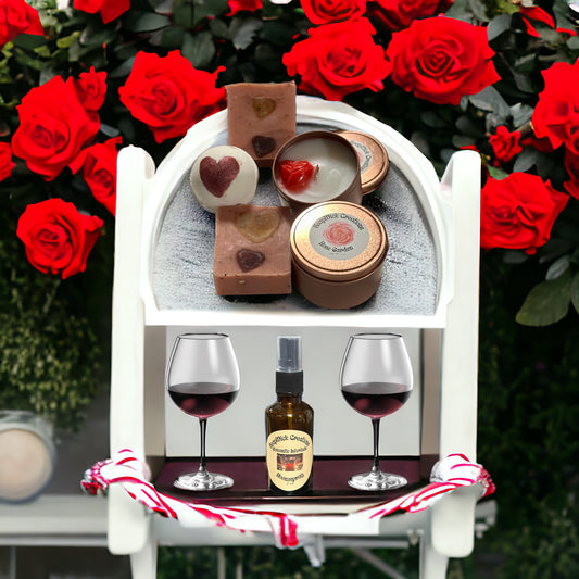 Photograph with a bar of rose scented soap, rose colored tin with a rose scented candle and a red rose embed in it, a Romantic Interlude bath bomb and Romantic Interlude Room spray on a white display case with two glasses of red wine with red roses in the background.