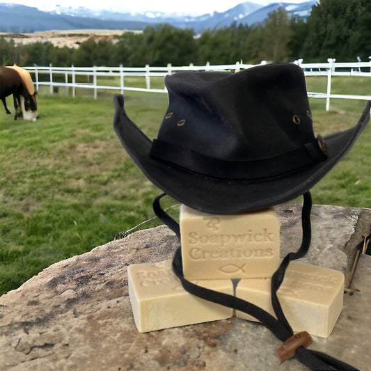 A photograph of cowboy clean soap with a cowboy hat sitting on top and a horse and horse ranch in the background.