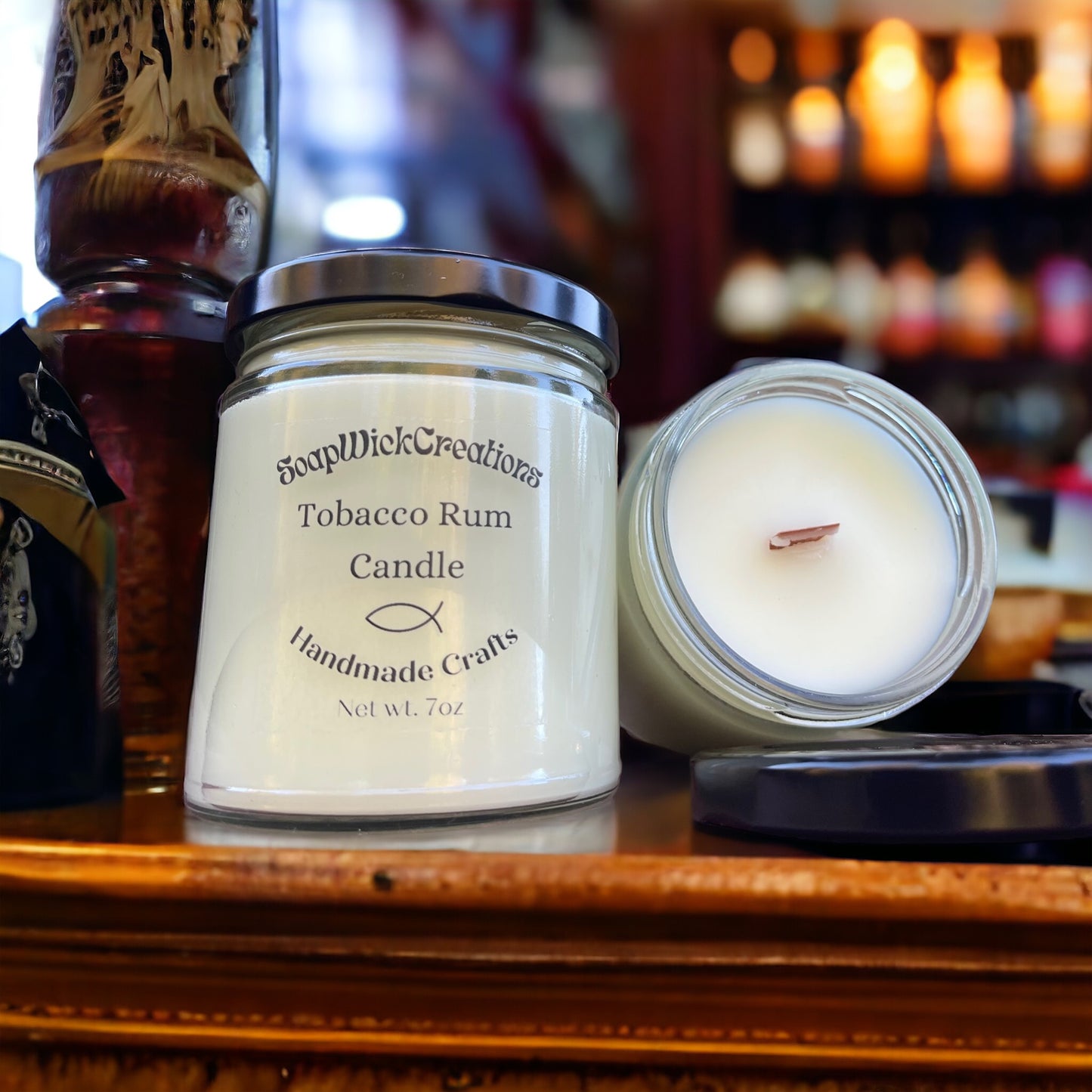 Two tobacco rum scented candles sitting on a bar-top with a wooden liquor bottle sitting next to them and bottles in a lit cabinet in the background.