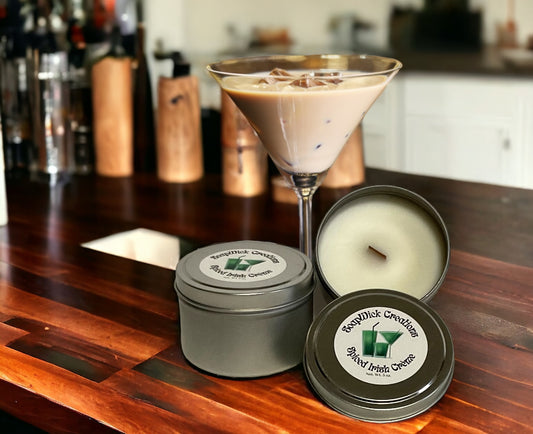 Two Spiced Irish Cream candles in metal tins with wooden wicks and a spiced Irish cream drink on a bar top and bar back in the background.