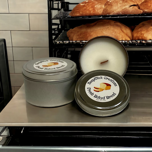 Three baked bread, white colored candles  with wooden wicks in metal tins on a stainless steel bakery  counter with loaves of baked bread in the background.