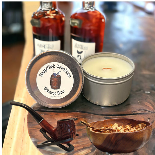 Tobacco Rum scented white candles in silver metal tins with two rum bottles in the background and a tobacco pipe, and a bowl full of tobacco in the foreground.