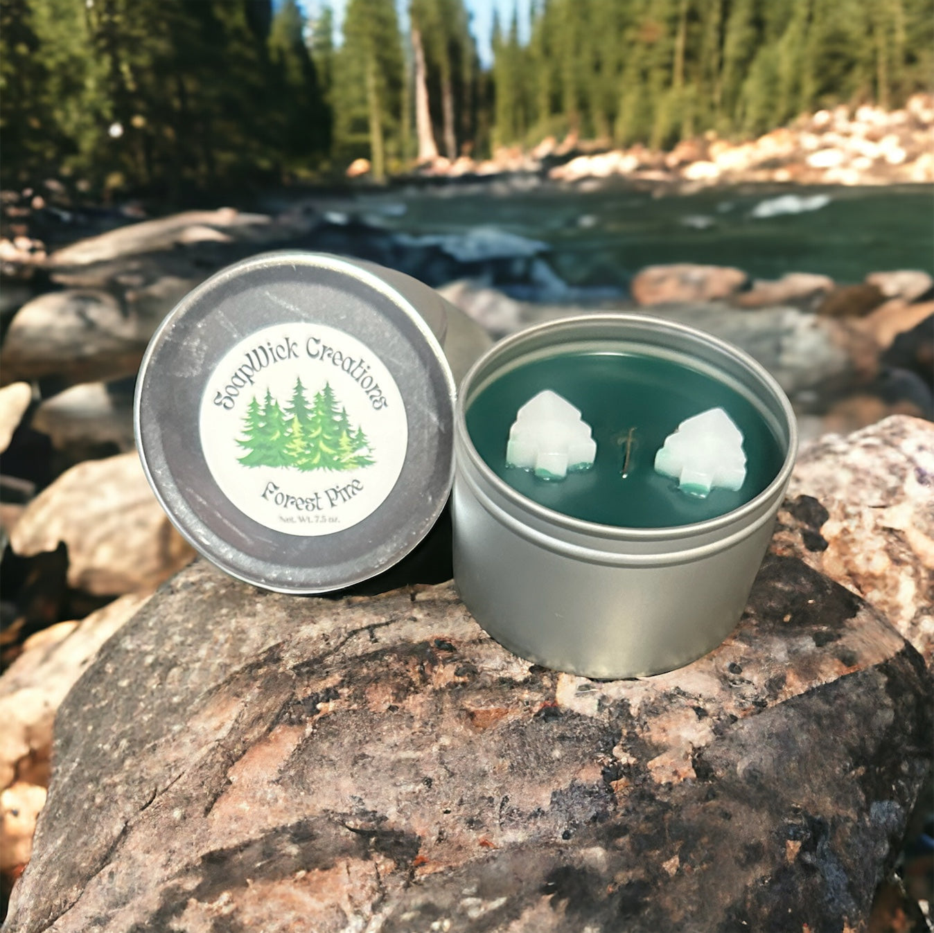 Forest Pine scented green candle in a silver metal tin with white pine tree embeds sitting on a rock with a river and pine forest in the background.