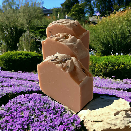 Three lavender scented bars of soap sitting on a rock wall with lavender, bushes and buildings in the background