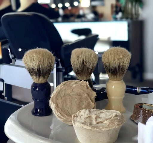Photograph of two dual lie, shaving pucks with shave brushes, and a barbers chair in the background in a barbershop.