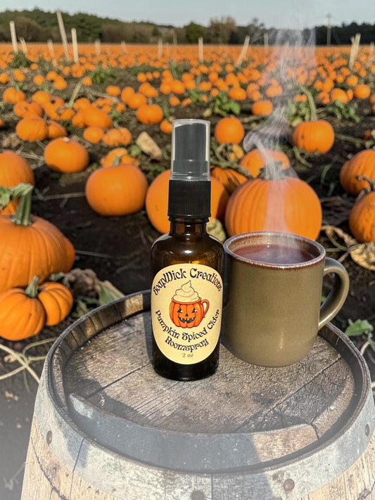 Amber bottle of Pumpkin Spiced Cider Room Spray sitting on an old apple barrel next to a steaming hot cup of apple cider with a pumpkin patch full of pumpkins in the background.