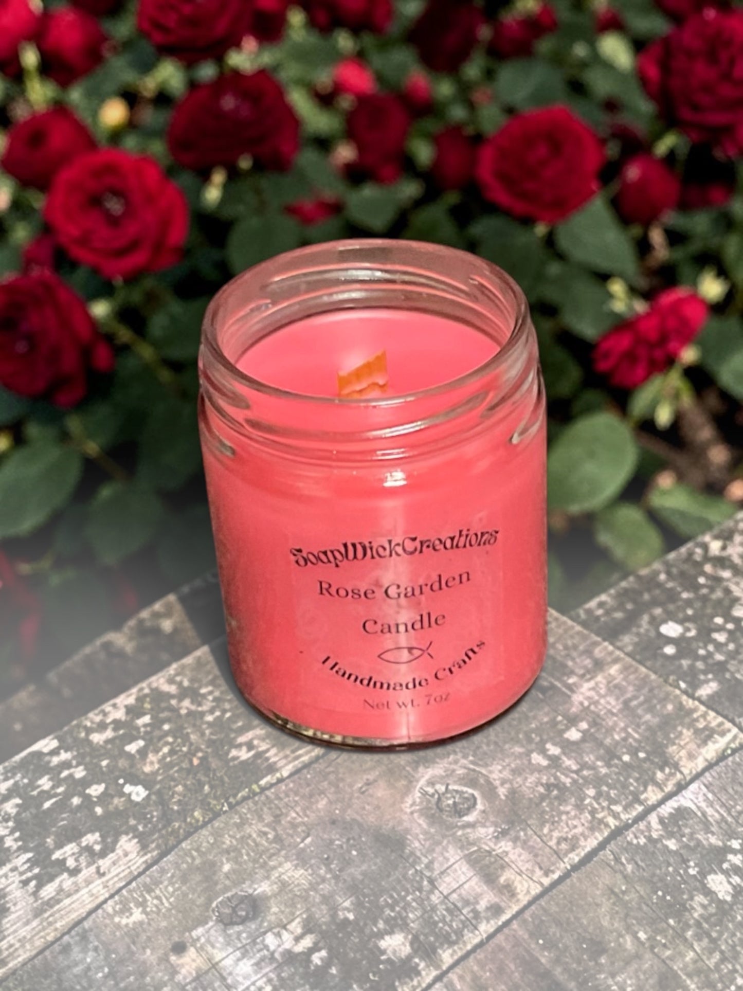 One red rose scented candle on a table sitting in a rose garden, surrounded by red roses.