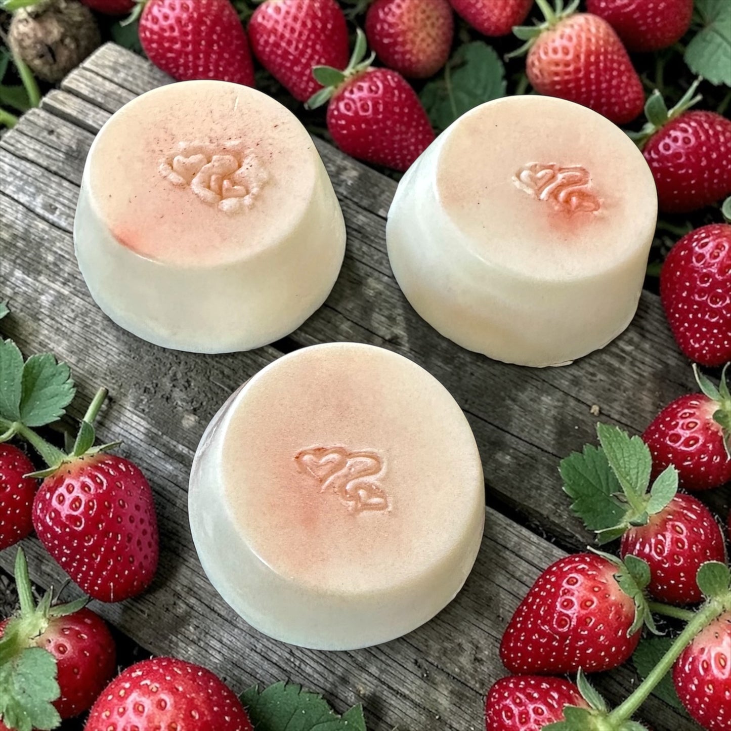 Three strawberry scented shampoo bars, sitting on a wooden pallet with strawberries in the background.