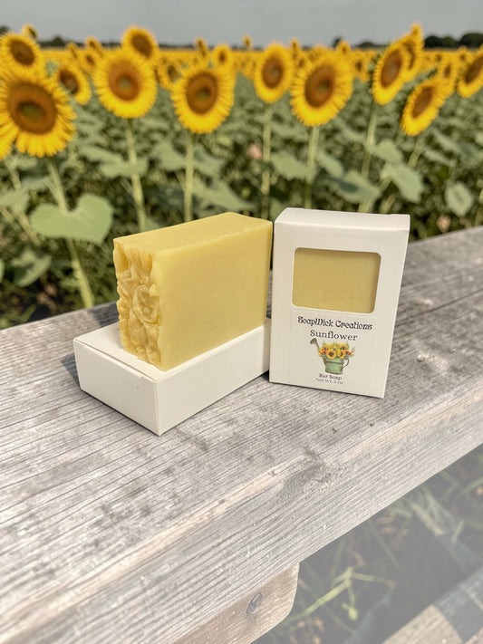 Two bars of yellow colored, sunflower, scented soap sitting on a wooden bench in a field of sunflowers.