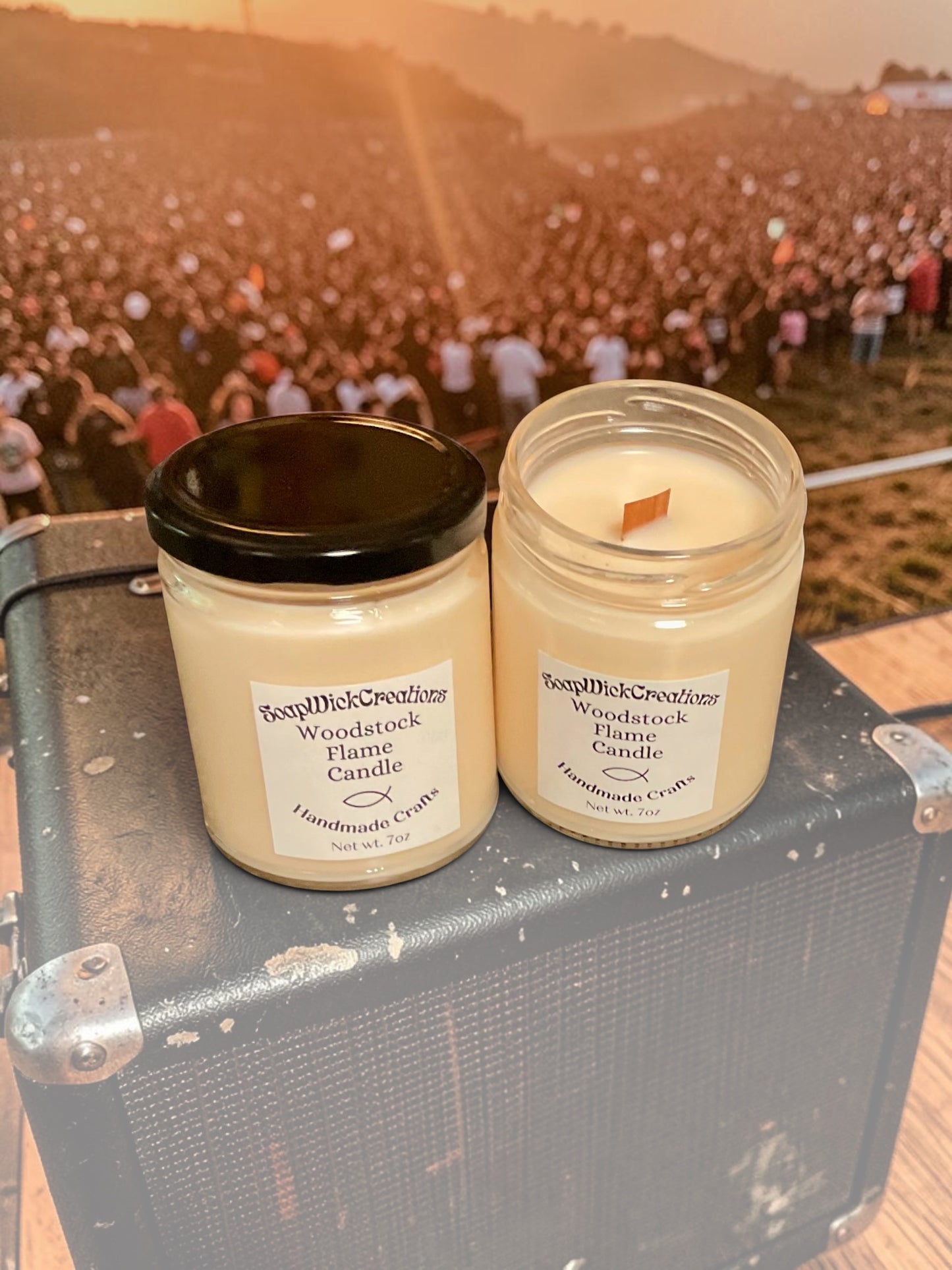 Two coconut candles in glass jars with cream-colored wax and white labels reading 'SoapWick Creations-Woodstock Flame Candle Handmade Craft' sitting on top of an old guitar amplifier with a blurred outdoor concert crowd in the background.