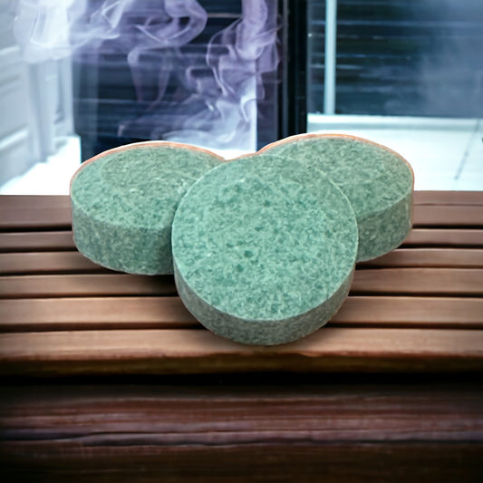 Three light green shower steamer tablets sitting on a bench inside a sauna with steam in the background.