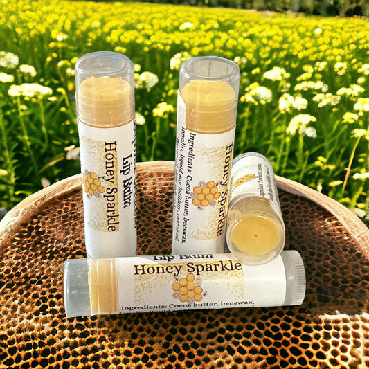 Four tubes of honey flavored Honey Sparkle lip balm, sitting on honeycomb with a clover-field in the background.