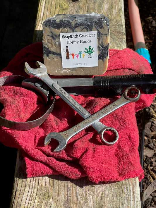 Hoppy Hands black and tan bar soap with pumice sitting on a board and shop rag with two combination wrenches and an oil filter wrench.