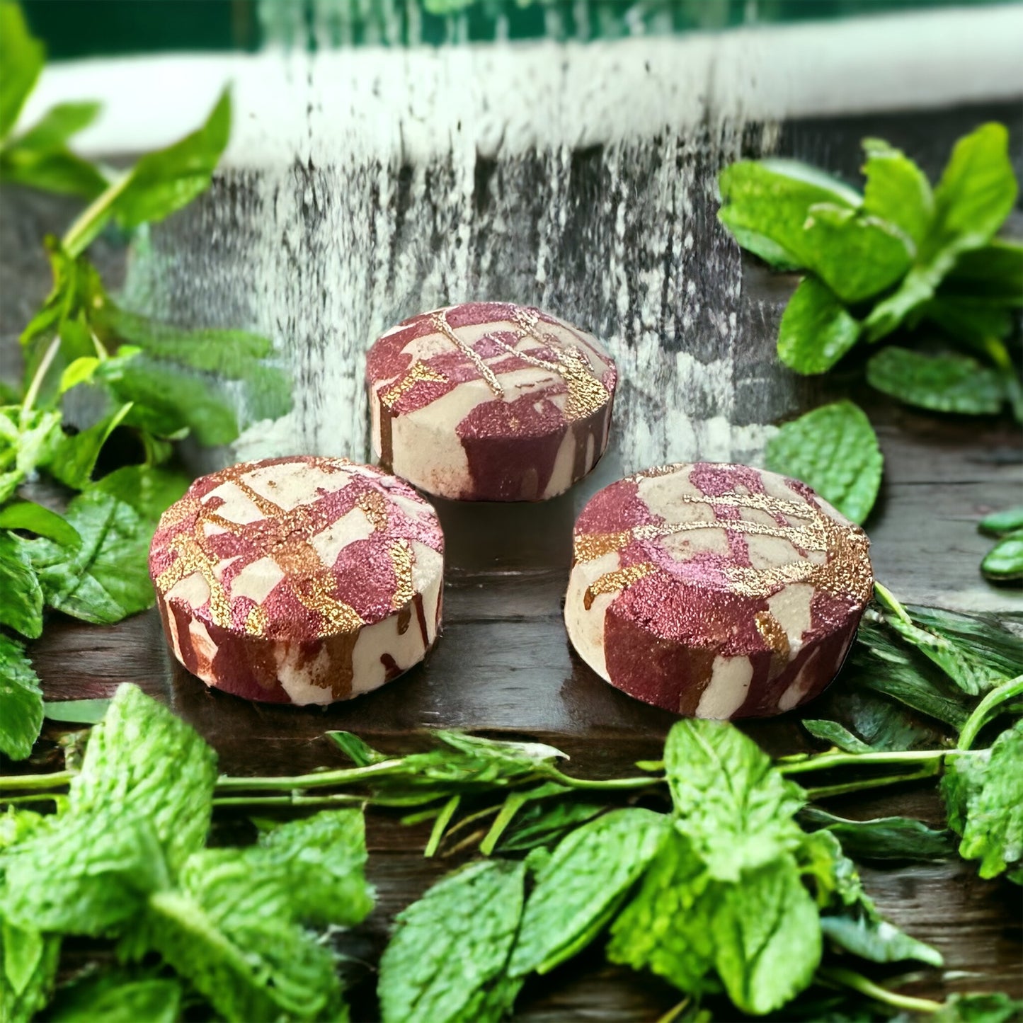 Photograph of three shower, steamers, maroon and white in color with mint and rosemary leaves around them underneath a waterfall.