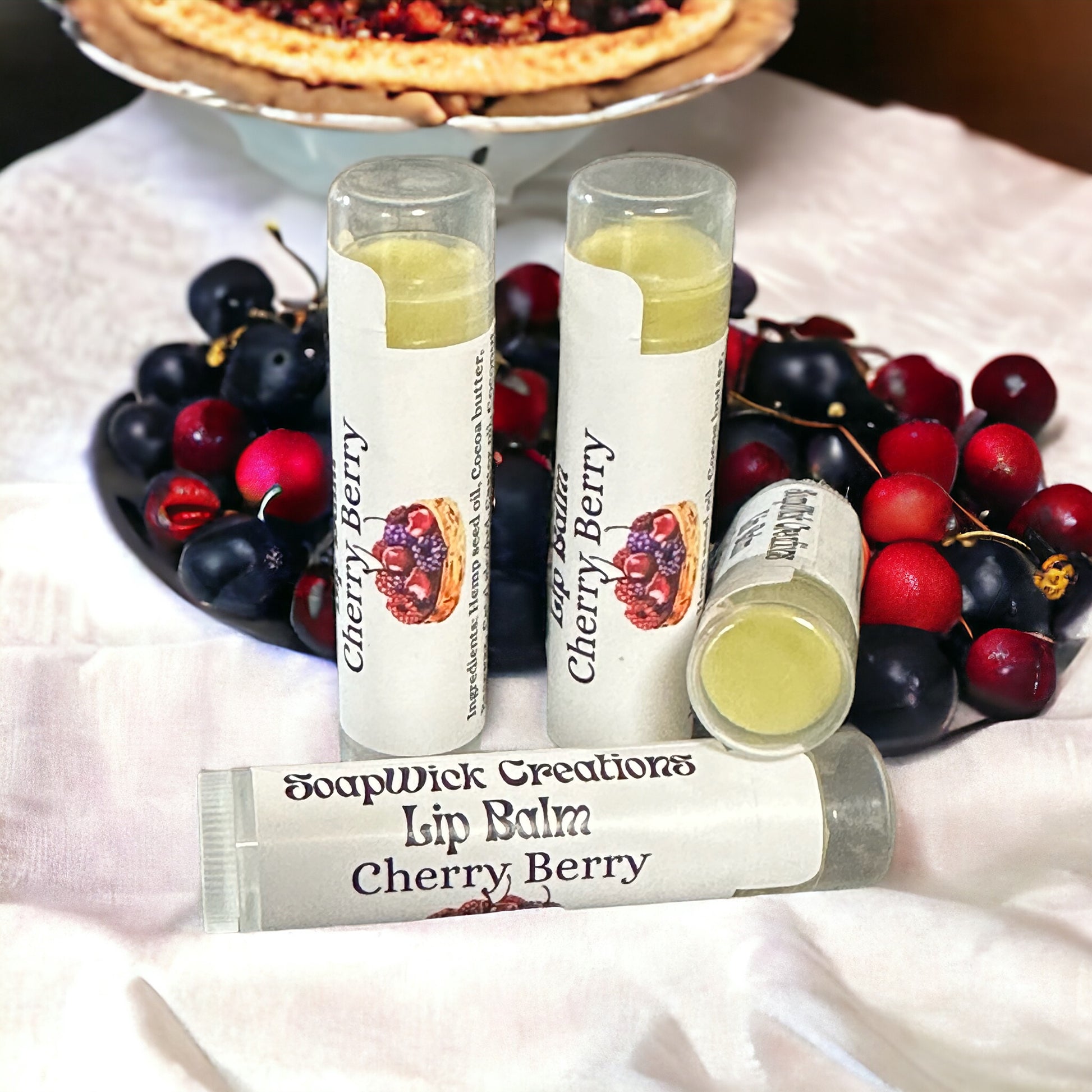 Four tubes of Cherry Berry lip balm next to a bowl of cherries and berries, with a pie in the background.