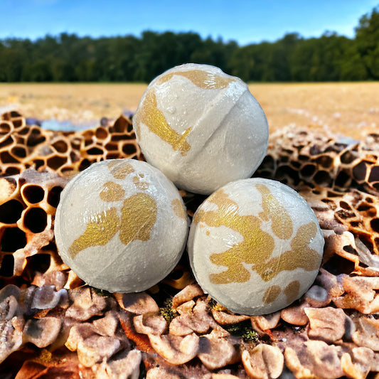 Three white with gold Honey and Oatmeal scented Bath Bombs, sitting on a honeycomb with oats and an oat field in the background.